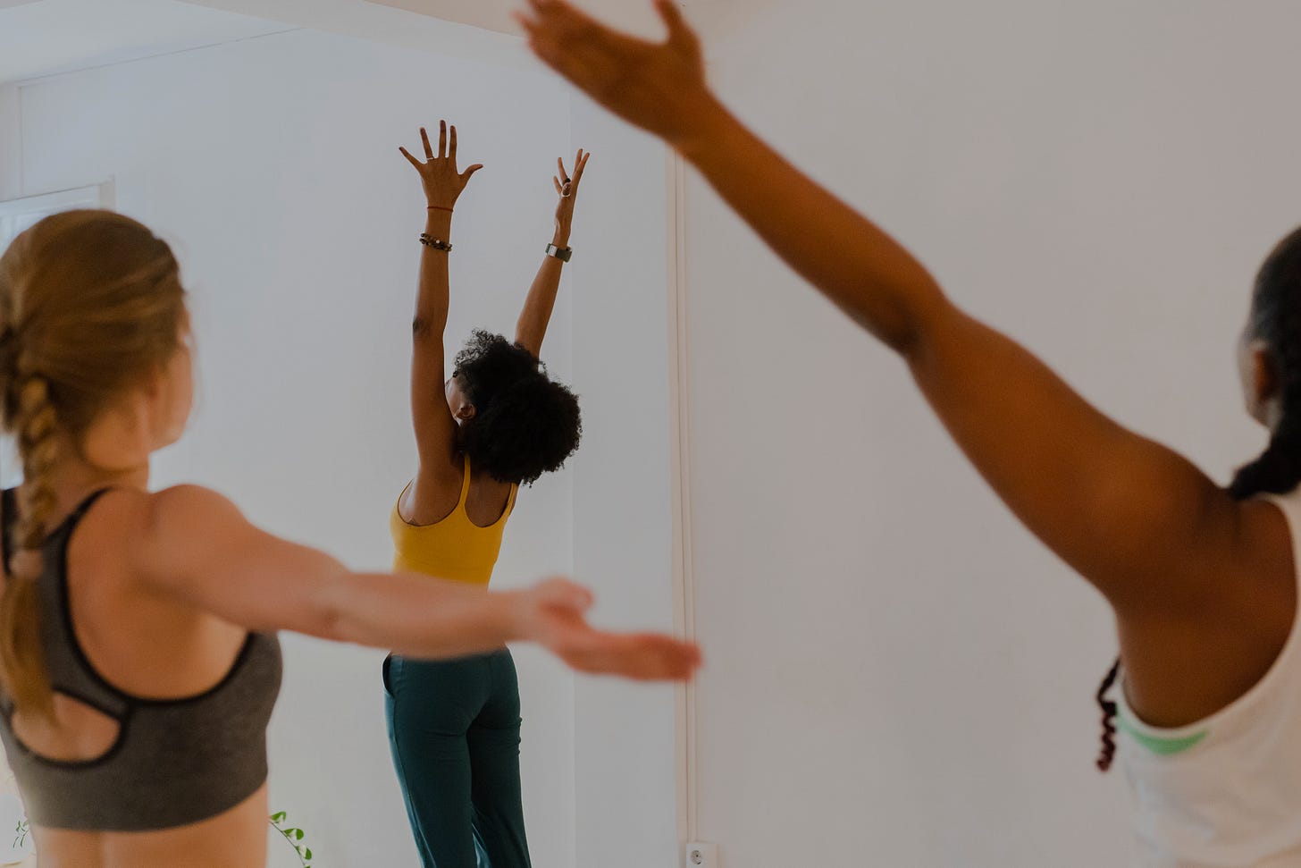 Three women practicing yoga. They are standing in mountain pose and reaching their arms up, we see them from the back.