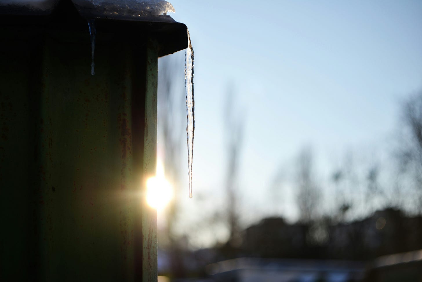 An icicle hangs from the edge of a structure, backlit by a low winter sun against a clear blue sky, with bare trees blurred in the background.