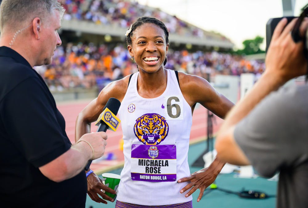 Michaela Rose wears a white LSU uniform and a bib with her name. She smiles at the camera as she talks to a reporter holding a microphone after her NCAA 800m title. Michaela Rose wears a white LSU uniform and a bib with her name. She smiles at the camera as she talks to a reporter holding a microphone after her NCAA 800m title.