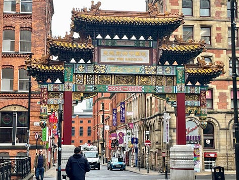 A mural showing a corner building in blue and green with the name 'SNOBS' written in white on its signage; a bright red and green and yellow archway with the words 'China Town Manchester' written on signage, with cars passing underneath; a mural showing a building with archways and 'MR EGG' signage in yellow, with a white car in front