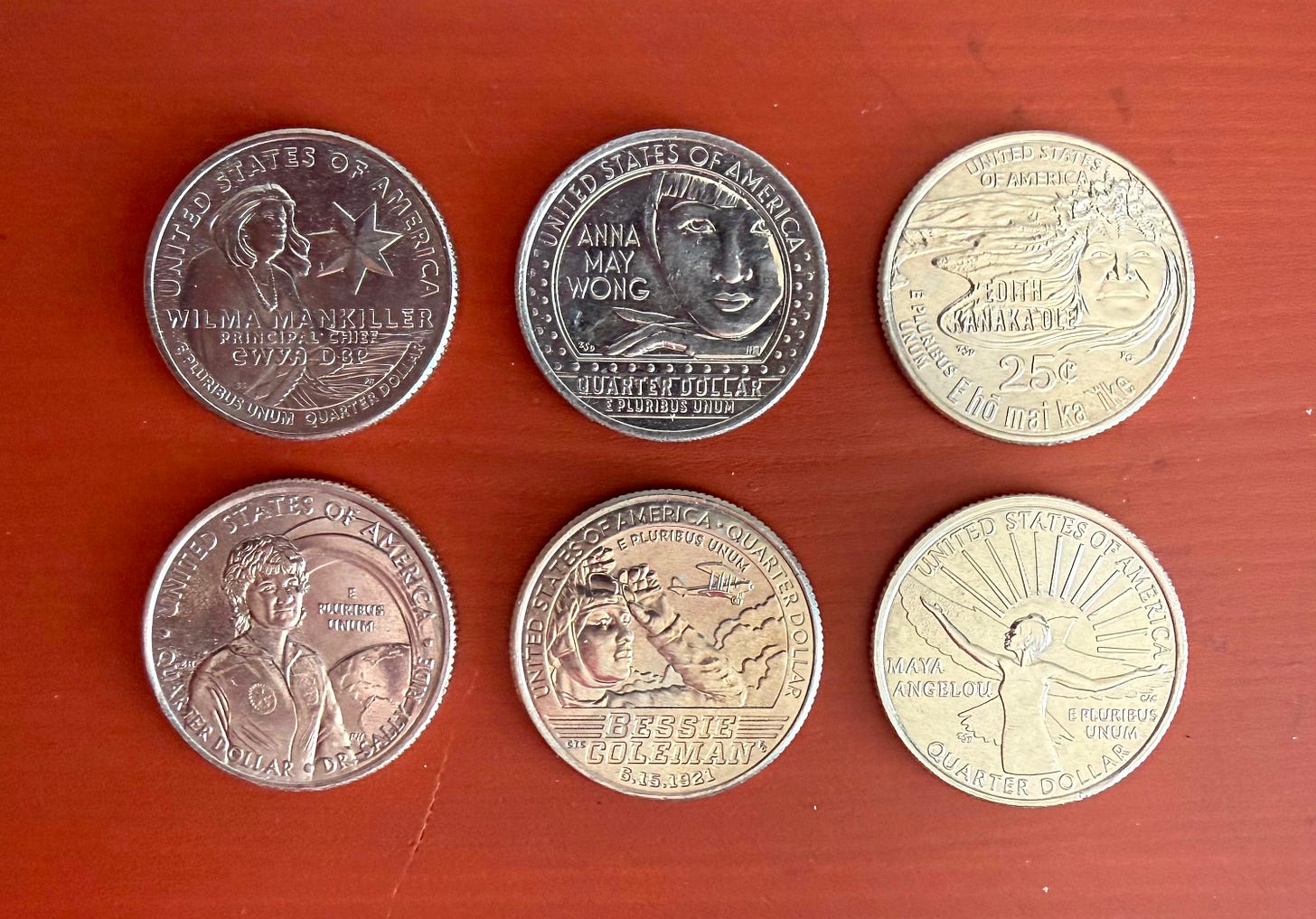 An array of the reverse sides of six U.S. quarters, arranged in two rows of three on a wooden surface painted reddish-brown. The coins are from the American Women Quarters Program.