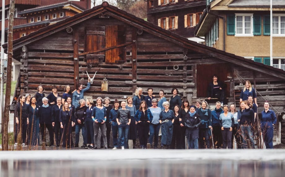 A group photo of 39 members of yodelling choir Echo vom Eierstock. The members are all women dressed in various shades of blue, standing in front of a Swiss alpine building.