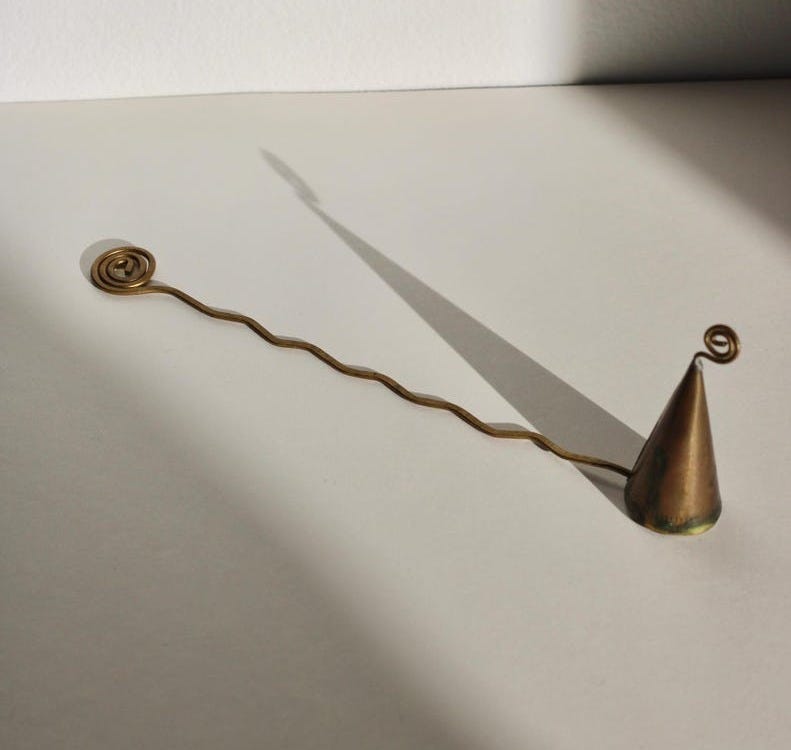 May include: A brass-colored metal incense holder. It features a cone-shaped incense holder with a spiral design at the top. A wavy line extends from the cone, ending in a spiral. The holder is on a white surface, with a shadow cast by sunlight.
