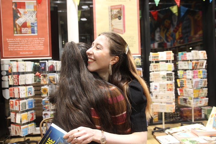 Author Isabelle Kenyon chats to two writers with a book and pen in her hand in a colourful bookshop (Blackwell's); In the second image author Isabelle hugs a reader with book 'The Dark Within Them' in her hand, in background the bookshop is selling cards on a portrait colourful rack.