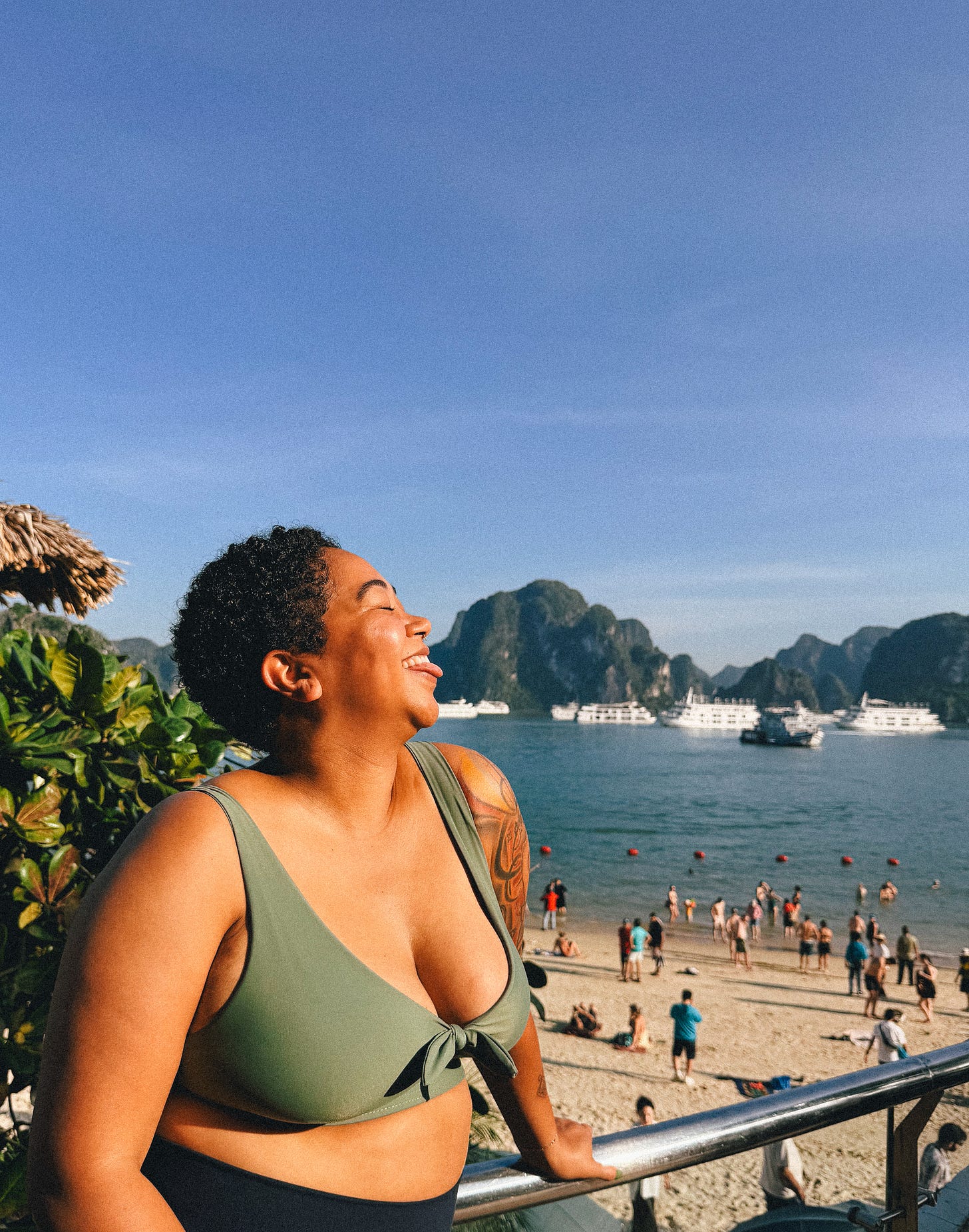 Anela stands in a bikini top smiling into the sun, while a beach pebbled with people is behind her and blue sky and water in the distance.