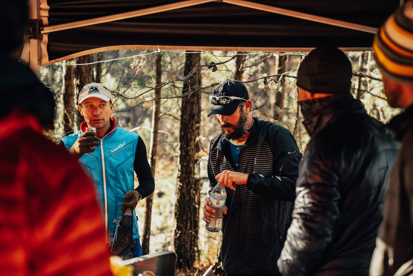 Two racers, one drinking a small can of something and the other opening a water bottle, and a group of aid station volunteers.