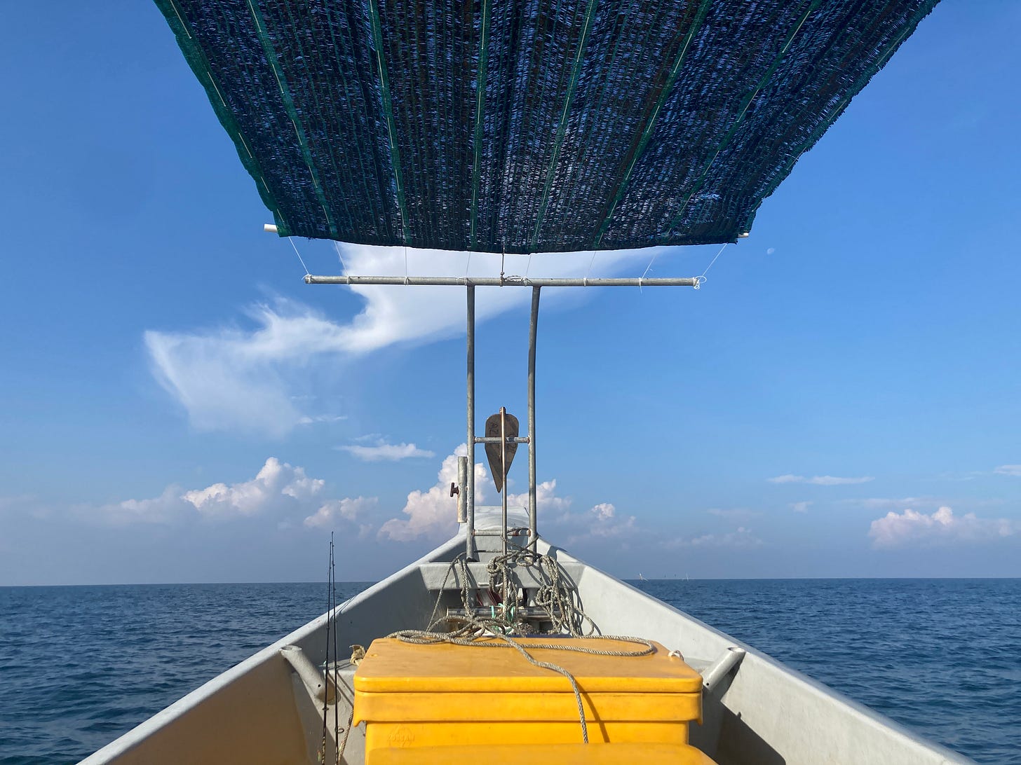 A picture of a prow of a ship bisecting a view of the sky and sea. Bright yellow cooler boxes stand in the foreground, and overhead, a thin sheet tries and fails to blot out the sun.