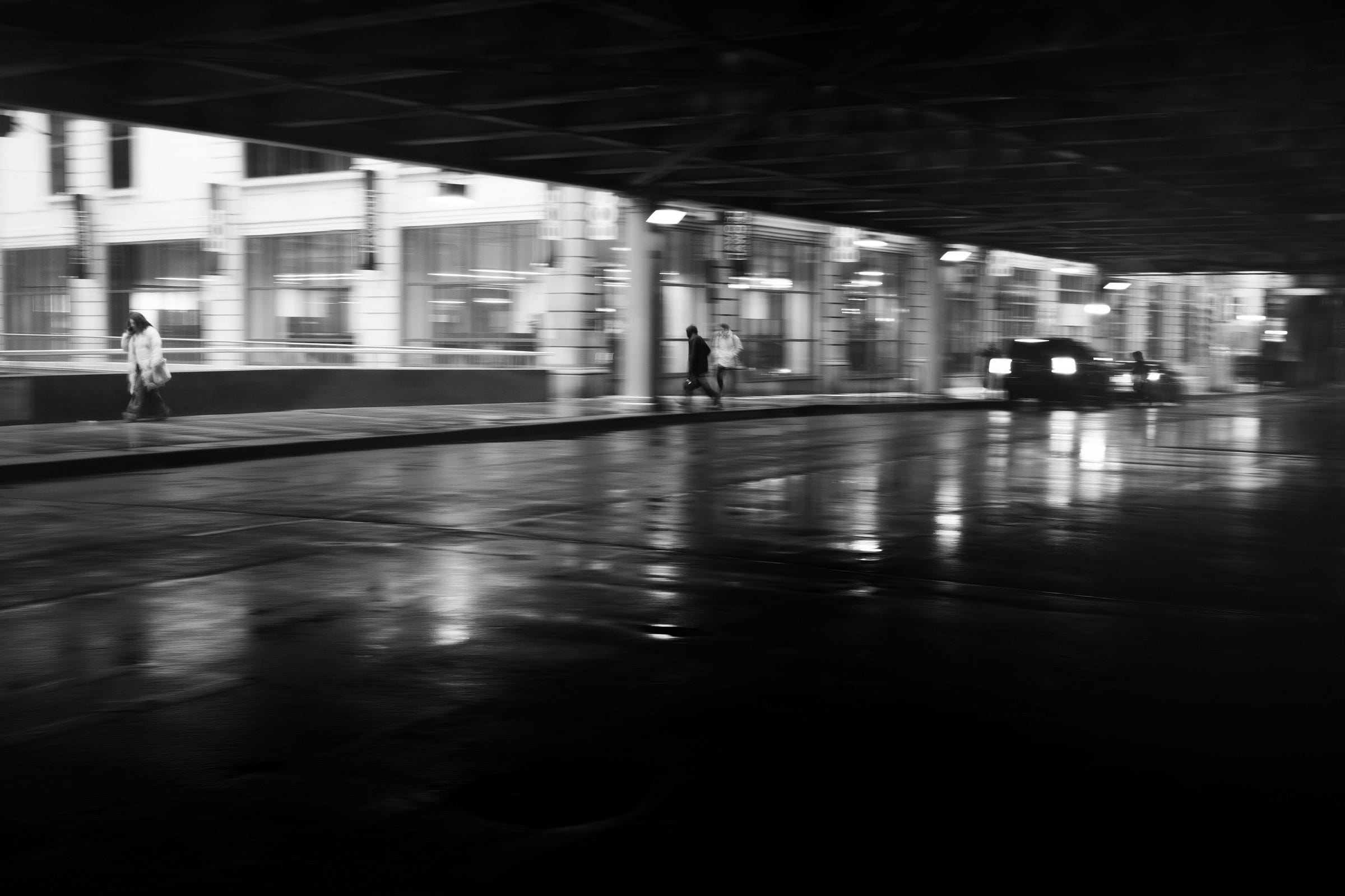 Blurry figures walk along a rain-soaked street under an overpass, with reflections shimmering on the wet pavement and a car's taillights glowing in the distance. Blurry figures walk along a rain-soaked street under an overpass, with reflections shimmering on the wet pavement and a car's taillights glowing in the distance.