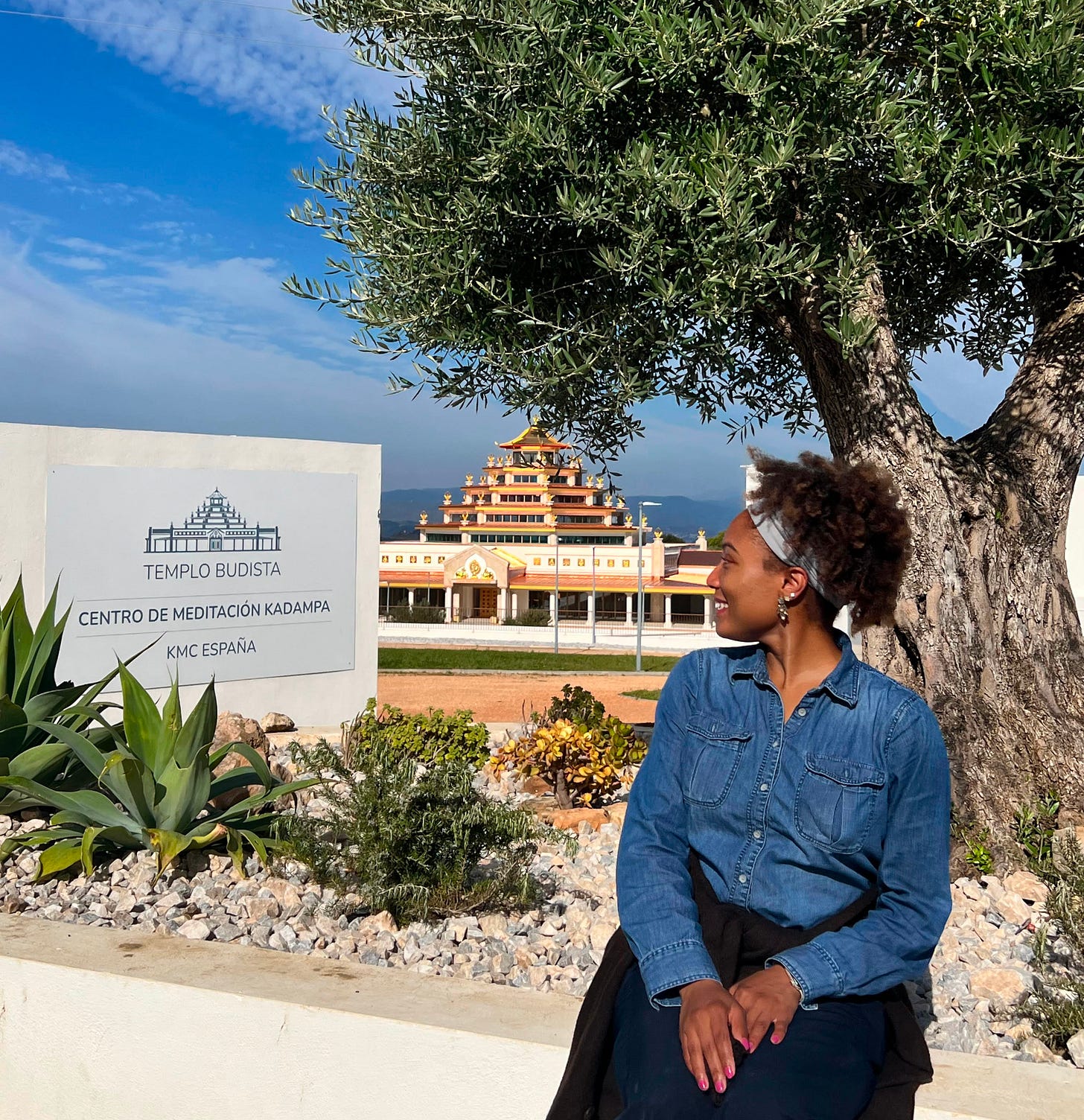 The author is sitting by the Templo Budista Centro de Meditación Kadampa KMC España sign, looking contemplatively toward the temple, with an olive tree and blue sky above. The author is sitting by the Templo Budista Centro de Meditación Kadampa KMC España sign, looking contemplatively toward the temple, with an olive tree and blue sky above.