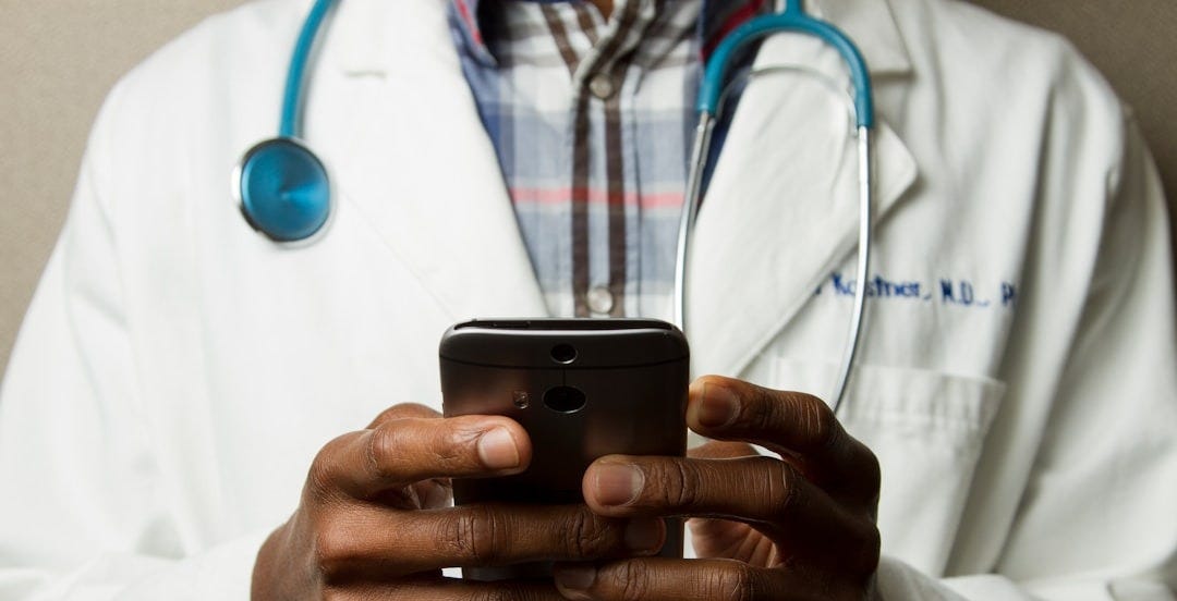 person wearing lavatory gown with green stethoscope on neck using phone while standing