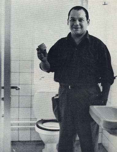 Piero Manzoni standing smiling in a toilet/bathroom with one of his tins of shit
