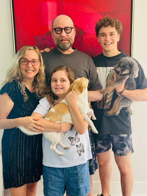 A collage of nine photos showing a family and their pets. Several images feature a smiling family of four—two adults and two children—posing together indoors, sometimes holding small dogs. One photo shows a child hugging an adult at a table in a tender moment. Another shows two small dogs standing side by side on a wooden floor. There’s a close-up of an orange lizard on a light surface. One outdoor photo shows an adult and child sitting together in sunlight, looking at each other. Another indoor photo shows the family grouped together again, smiling. A selfie captures two adults close together, both wearing glasses. One image shows an adult sitting in a car holding a small dog wrapped in a blanket. The final photo shows two small rodents on a pink surface, facing the camera.