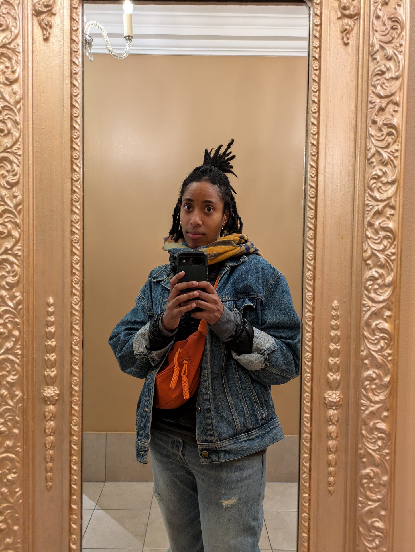 Photo of Natalie wearing a denim jacket, jeans, a colorful scarf and red crossbody bag taking a 3/4 body selfie in a gold-framed mirror. Photo of Natalie wearing a denim jacket, jeans, a colorful scarf and red crossbody bag taking a 3/4 body selfie in a gold-framed mirror.