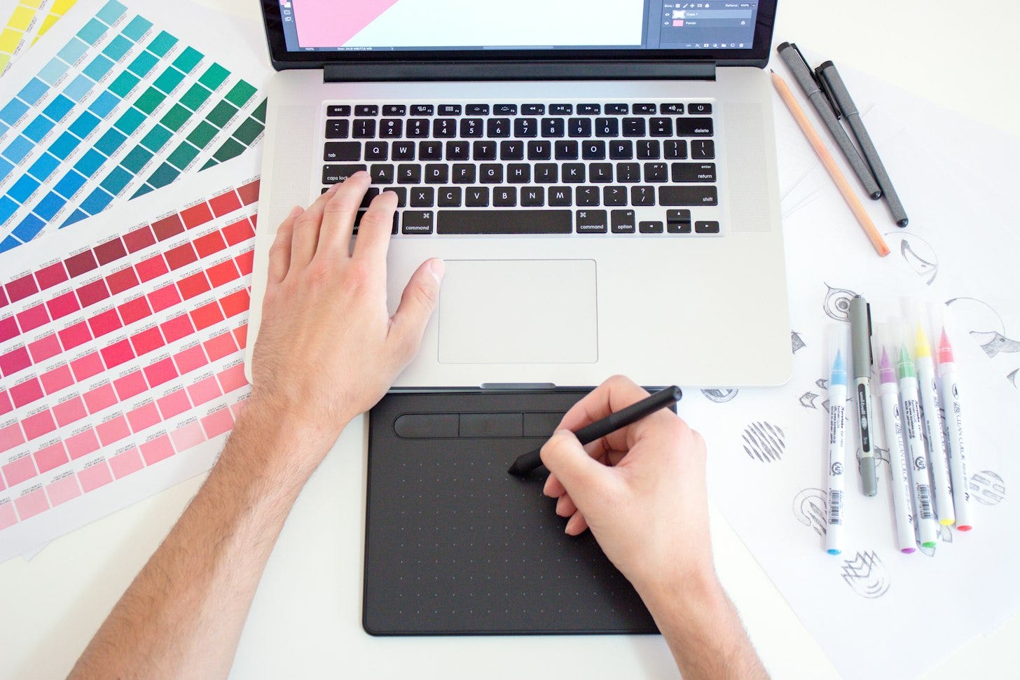 Overhead view of a person’s hands on a laptop and a drawing tablet, beside printed color swatch cards and a row of markers on a white desk. Overhead view of a person’s hands on a laptop and a drawing tablet, beside printed color swatch cards and a row of markers on a white desk.