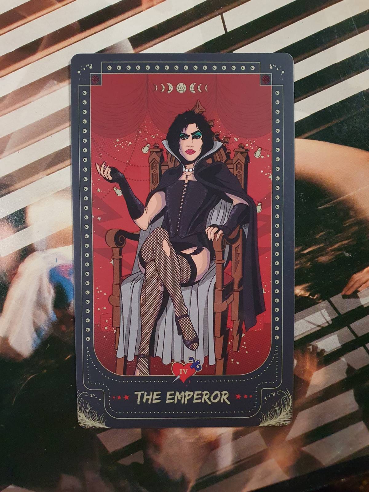 The Emperor tarot card featuring a dark-haired gothic figure in a black cape, corset and fishnet stockings, seated confidently on a throne against a red theatrical backdrop. The Emperor tarot card featuring a dark-haired gothic figure in a black cape, corset and fishnet stockings, seated confidently on a throne against a red theatrical backdrop.