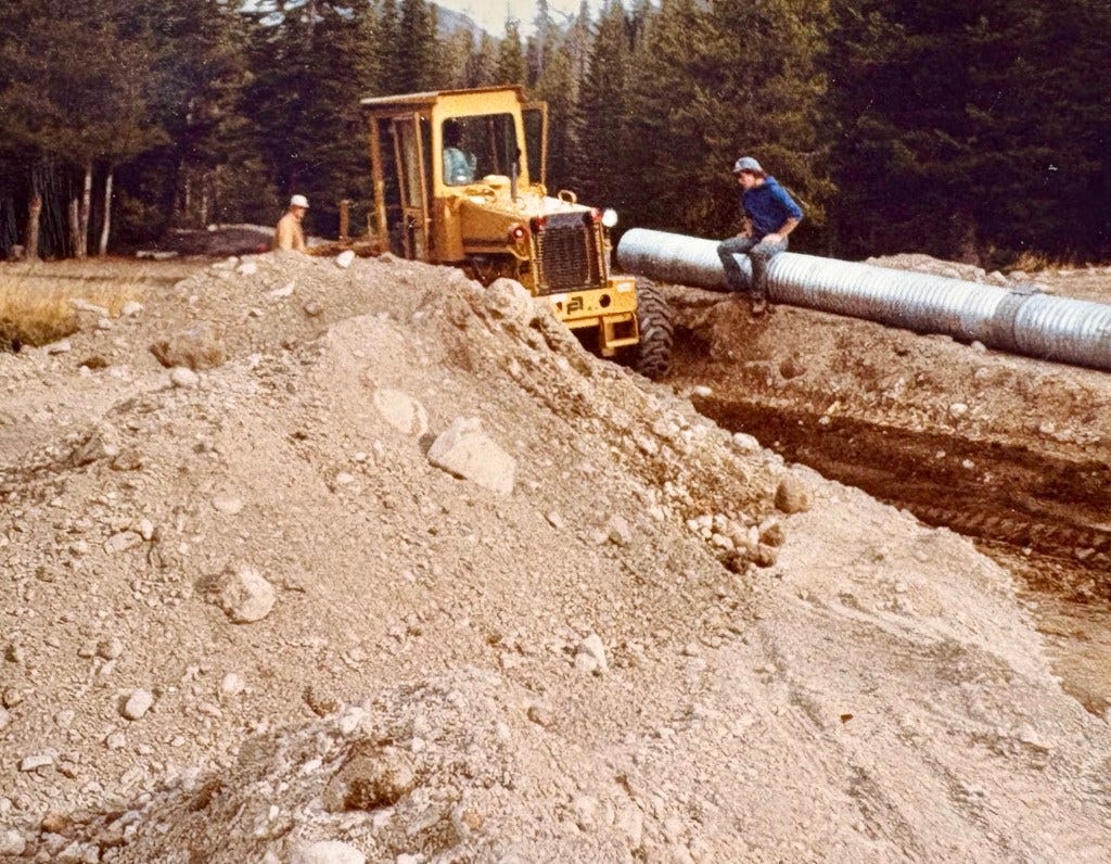 Two workers at a construction site, one on a bulldozer, with a mound of dirt and large metal piping in the foreground surrounded by trees. Two workers at a construction site, one on a bulldozer, with a mound of dirt and large metal piping in the foreground surrounded by trees.