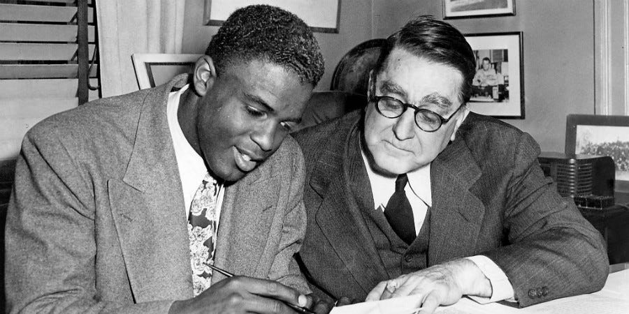 A black and white photograph of Jackie Robinson and Branch Rickey. Robinson appears to be signing his MLB contract.