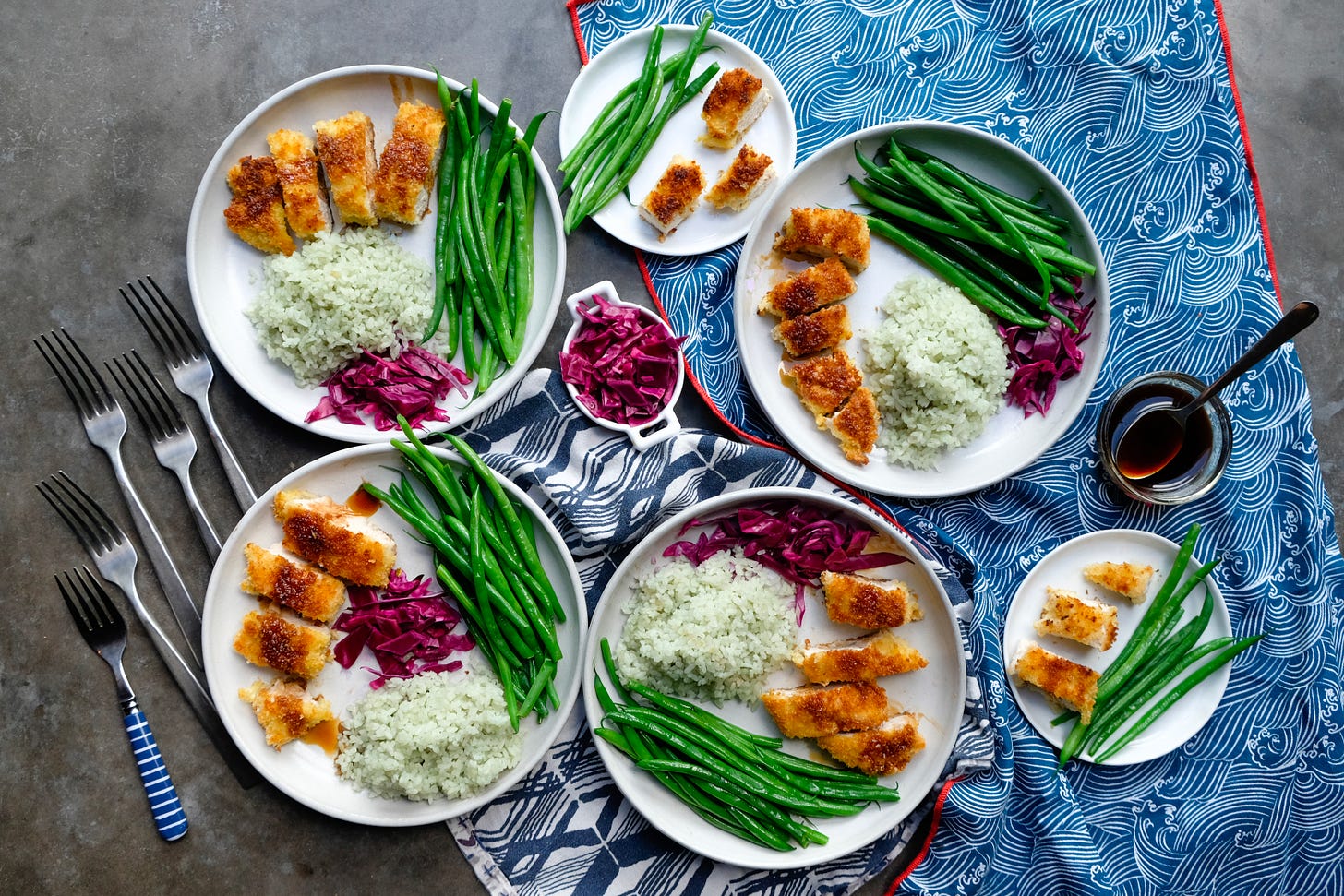 Five plates of Turkey Katsu (breaded turkey paillards) with jade rice, haricot vert, jade rice, and pickled cabbage, on a gray table with a blue tablecloth