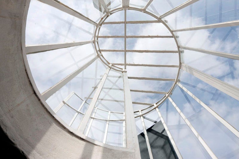 A view of a metal spiral staircase looking up from the bottom through glass to the clouds and sky.