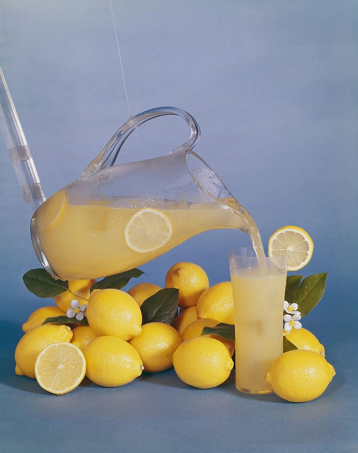 A glass pitcher pours lemonade into a glass, surrounded by fresh lemons and leaves on a blue background. A lemon slice decorates the glass.