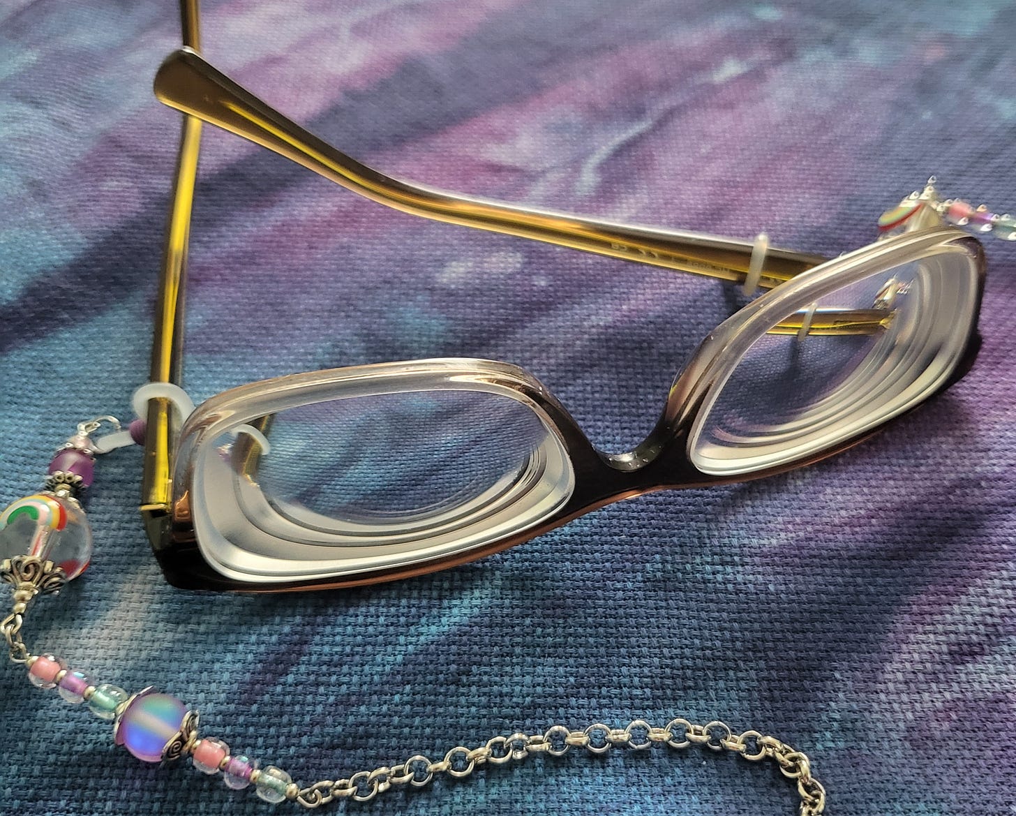 A pair of glasses with thick lenses and a rainbow chain lanyard rests on galaxy colored cross stitch fabric.