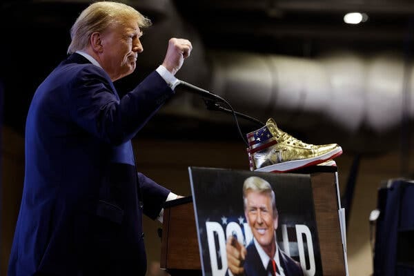 Former President Donald J. Trump stands onstage at a microphone. Before him on the podium is a gold hightop sneaker with an American flag at the ankle. Former President Donald J. Trump stands onstage at a microphone. Before him on the podium is a gold hightop sneaker with an American flag at the ankle.