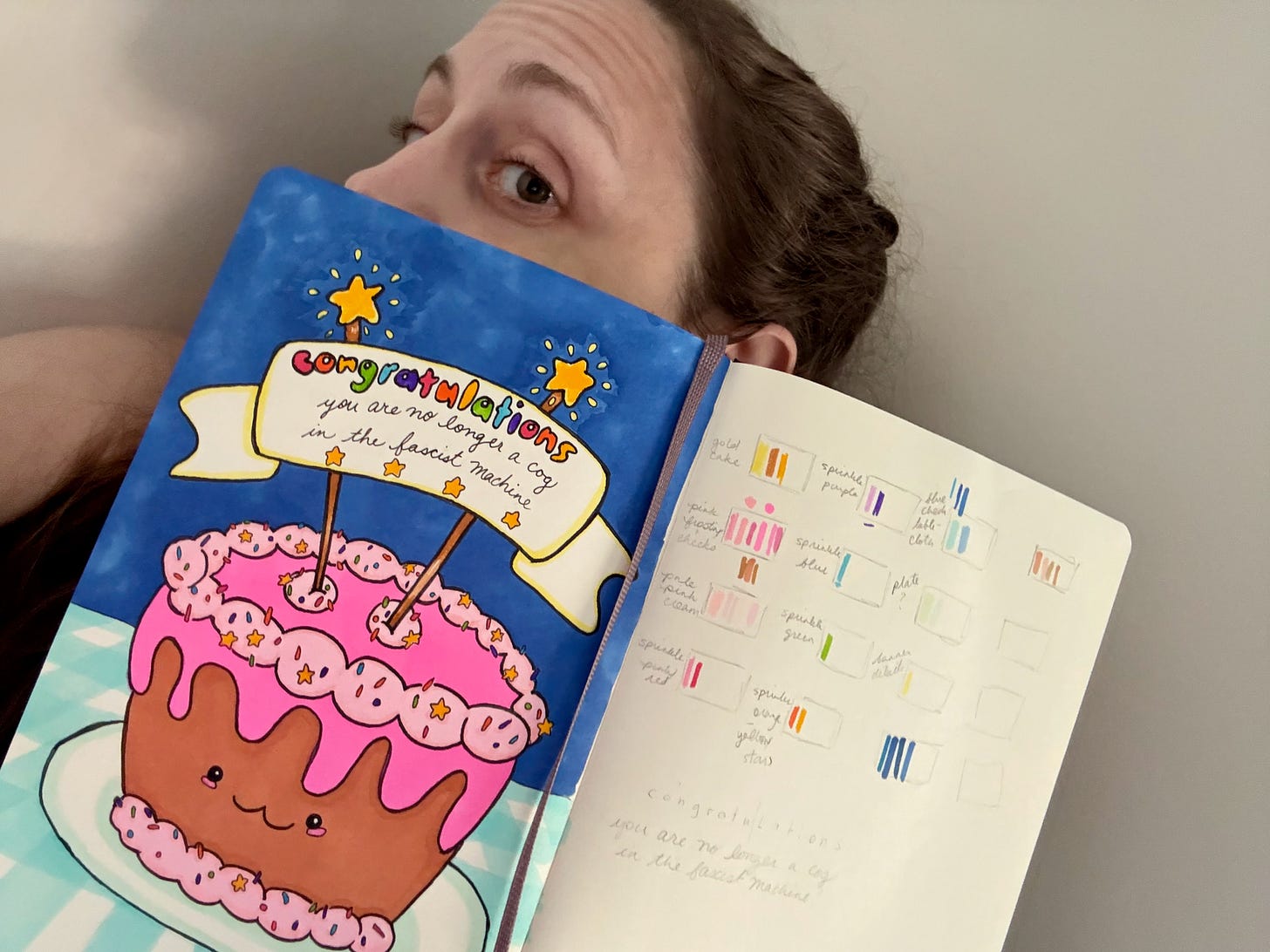 Selfie of Kate, with most of her face obscured by her sketchbook, open to a cartoon drawing of a happy little cake with a banner reading “congratulations / you are no longer a cog in the fascist machine”