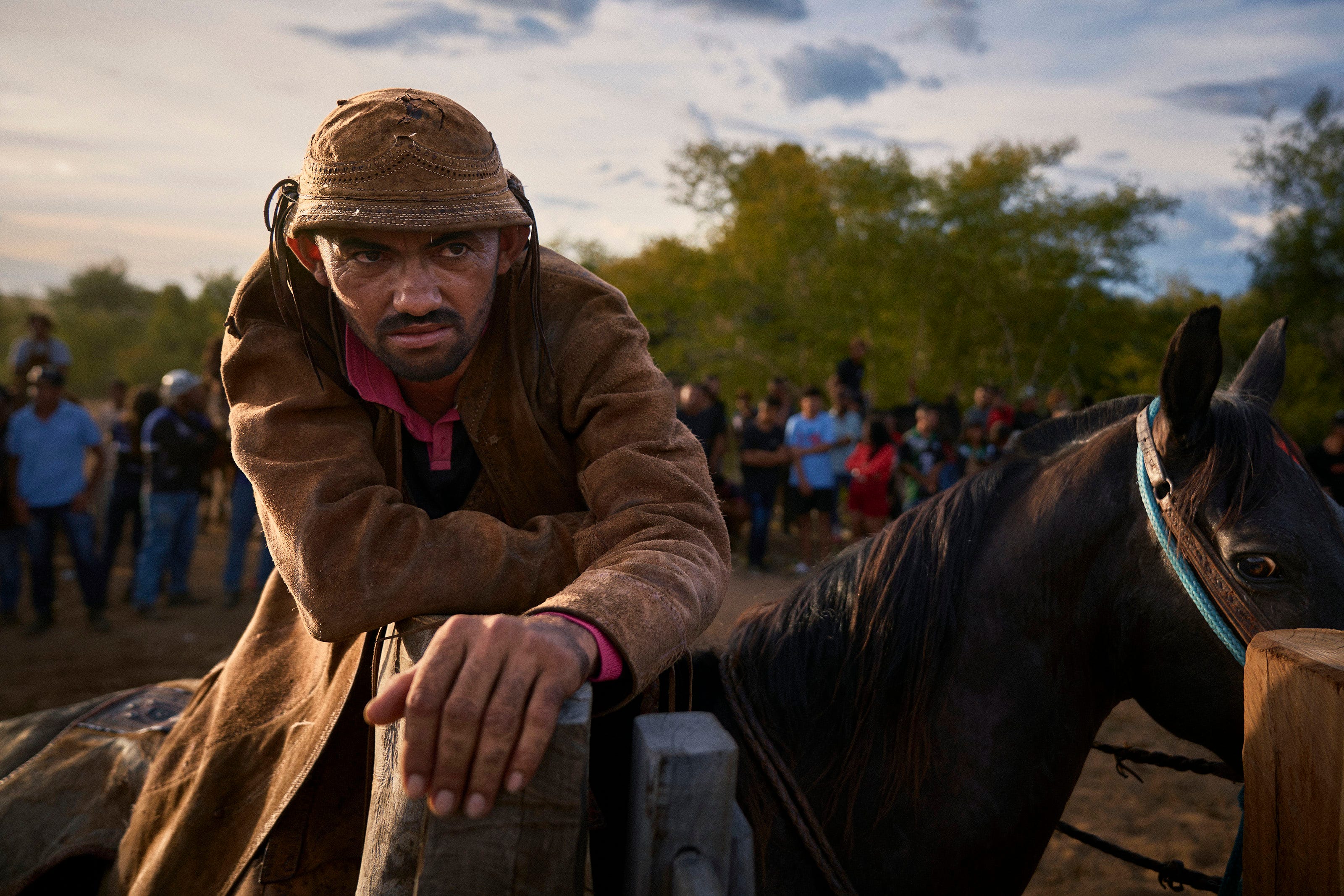 A man in a brown jacket and hat leans on a wooden fence beside a dark horse, with a crowd of people and trees visible in the background under a cloudy sky.