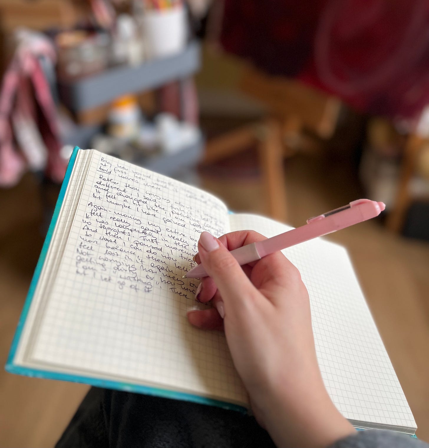 A close-up of a hand holding a pink pen, writing in a blue journal with a grid-patterned page. The background is softly blurred, showing an artist's studio with a rolling cart of supplies and an easel, creating a quiet, reflective moment within the creative space.
