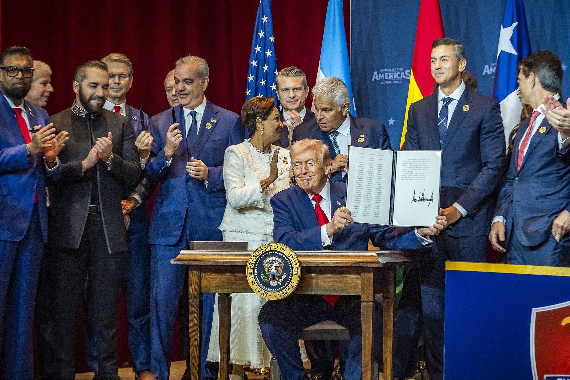 Donald Trump, seated at a desk bearing the presidential seal, holds up a signed document while about a dozen Latin American leaders in suits stand behind him applauding, in front of U.S., Argentine, Bolivian and Chilean flags. Donald Trump, seated at a desk bearing the presidential seal, holds up a signed document while about a dozen Latin American leaders in suits stand behind him applauding, in front of U.S., Argentine, Bolivian and Chilean flags.