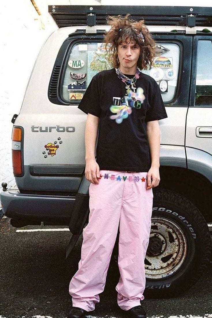 This may contain: a young man standing in front of a parked car wearing pink pants and a black t - shirt This may contain: a young man standing in front of a parked car wearing pink pants and a black t - shirt
