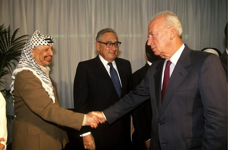 Three men stand, two shaking hands. Three men stand, two shaking hands.
