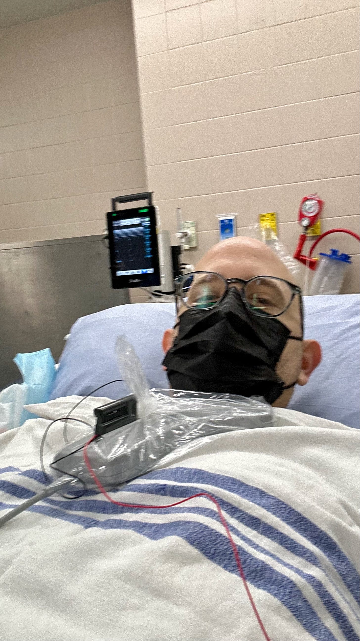 bald man with glasses wearing a mask under blankets on a hospital bed