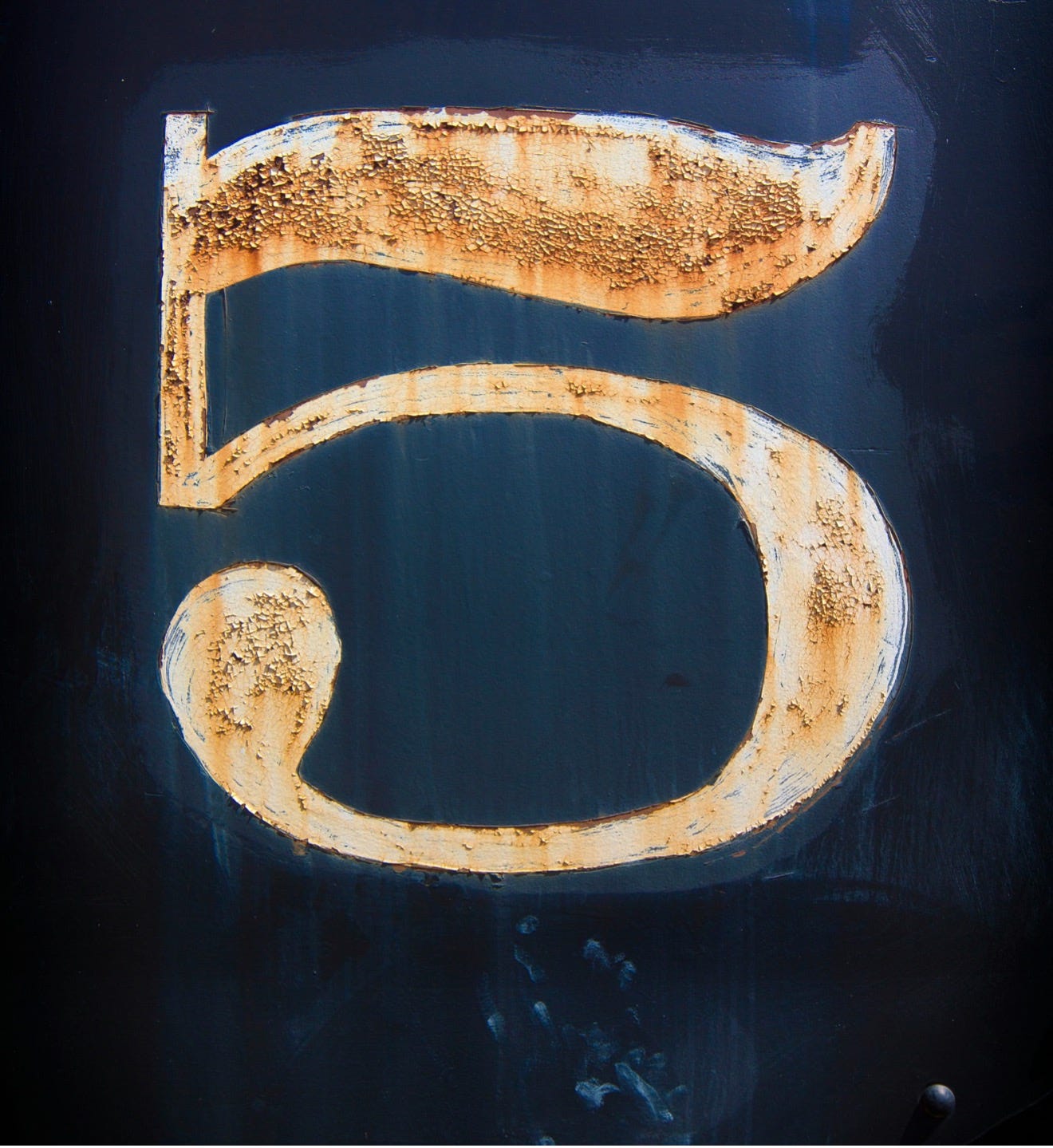 The number five painted in white on a dark metal background. Rust is showing through the paint.