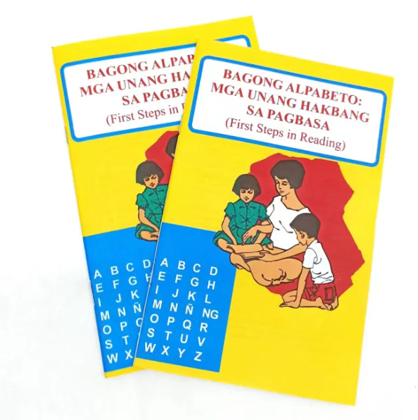 Two identical books on top of each other, titled 'Bagong Alpabeto: Mga Unang Hakbang sa Pagbasa.' The cover is bright yellow with the illustration of a woman reading to a boy and a girl. On the lower left corner of the cover is the Filipino alphabet. 
