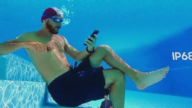 A man sitting at the bottom of a pool in swimwear and goggles looking at a Samsung phone