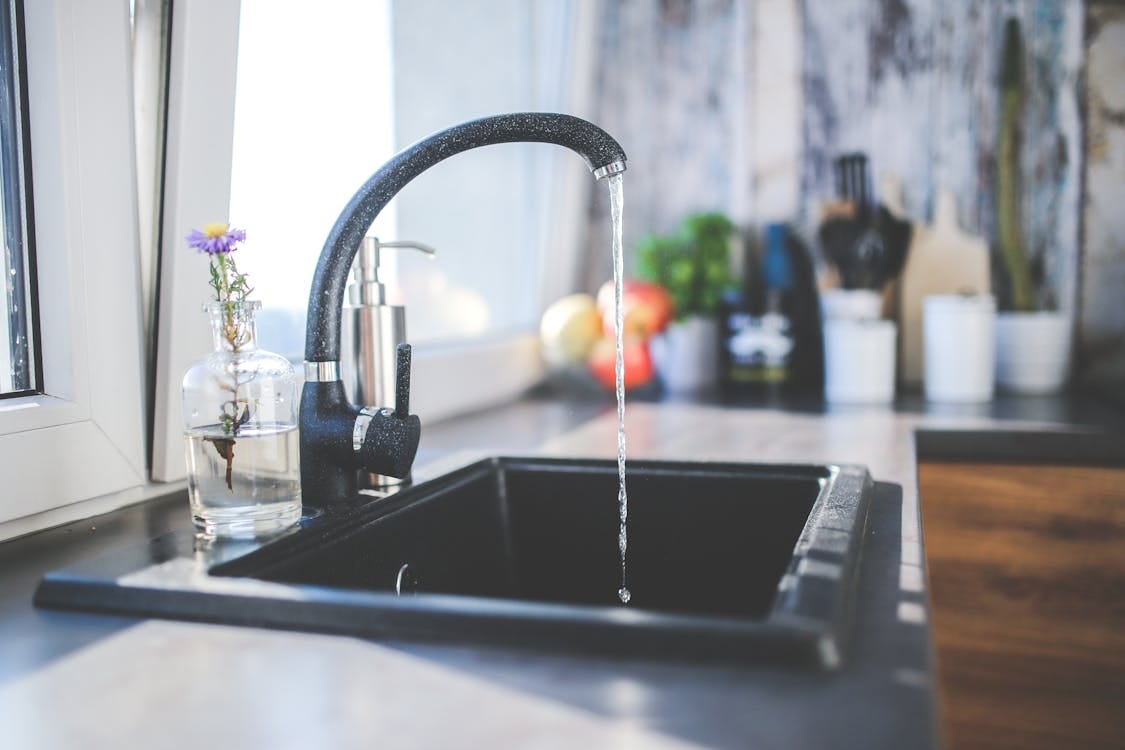 Free A modern kitchen sink with running water, a sleek faucet, and a vase on the countertop. Stock Photo