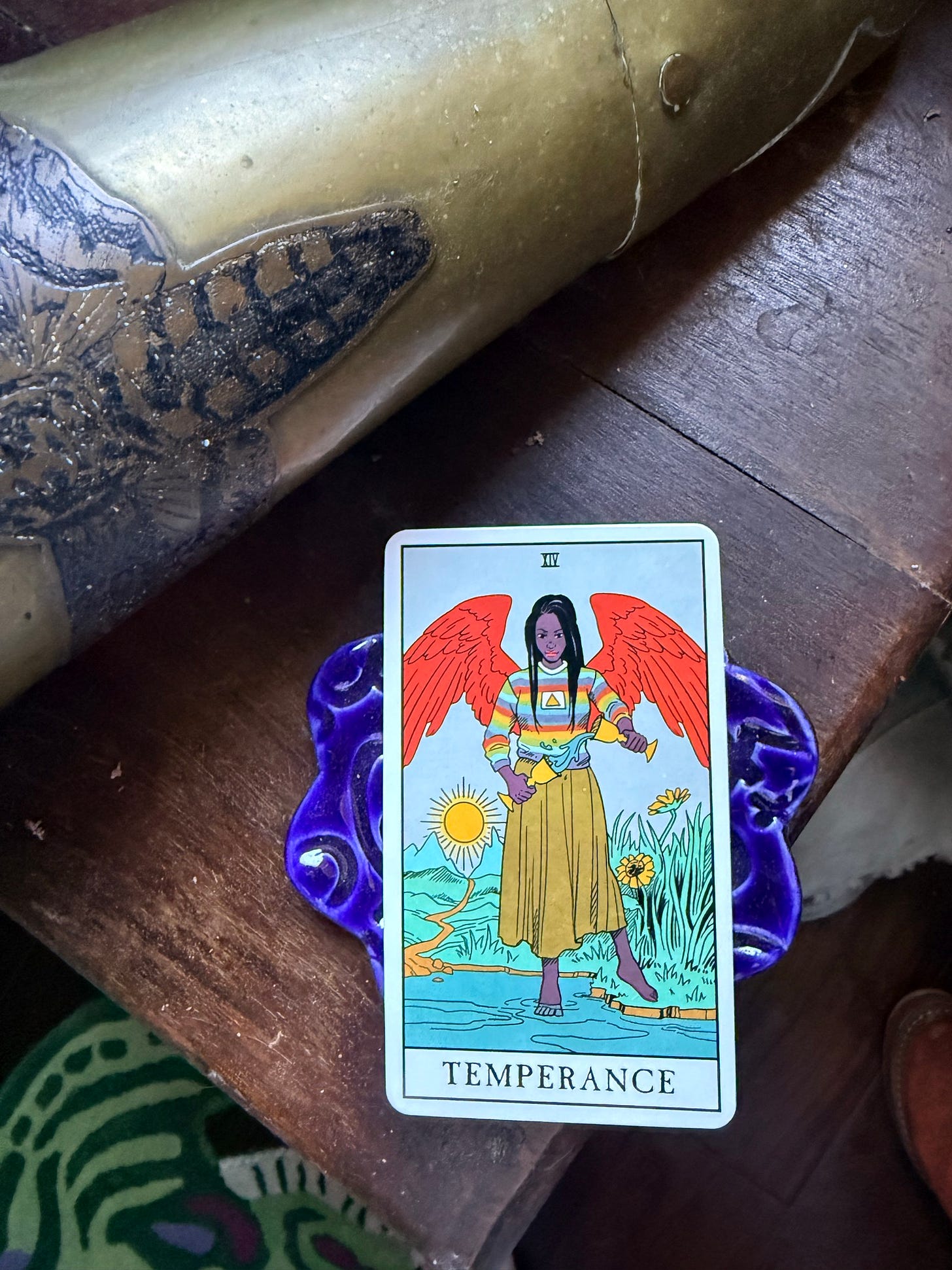 The Temperance card from the Modern Witch Tarot, depicting a red-winged figure pouring liquid between two golden cups, displayed on a purple ceramic stand beside a brass horn. The Temperance card from the Modern Witch Tarot, depicting a red-winged figure pouring liquid between two golden cups, displayed on a purple ceramic stand beside a brass horn.