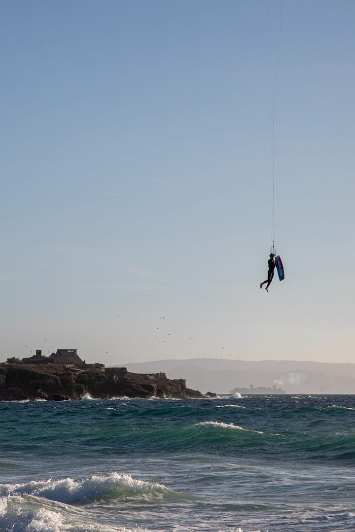 Kitesurfer riding near the medieval castle walls in Tarifa; the Mar Mediterráneo and Océano Atlántico signs marking where two seas meet at the Tarifa causeway; a kitesurfer suspended mid-air above the Strait of Gibraltar; a man running into the surf with a kiteboard on Tarifa beach