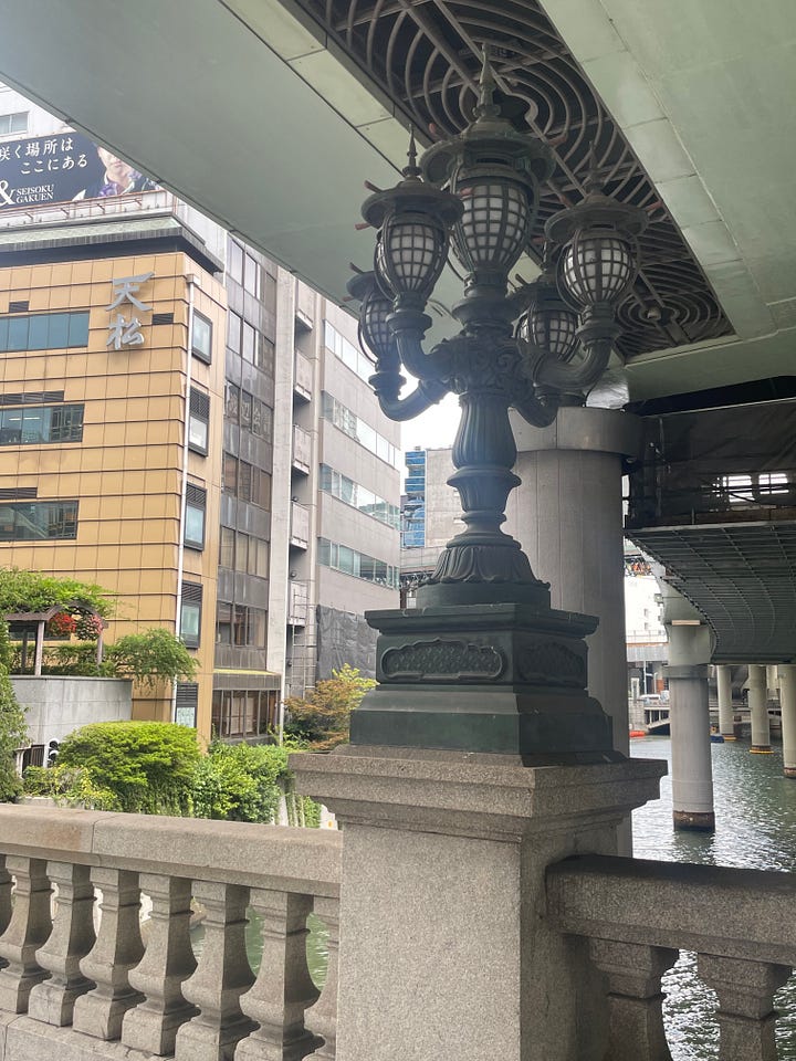 Photographs of ornate metal lanterns standing on Nihonbashi Bridge, one of which is tall and stands between the two flyover structures above, and the other is shorter and is seen with only light green metalwork above it instead of sky.