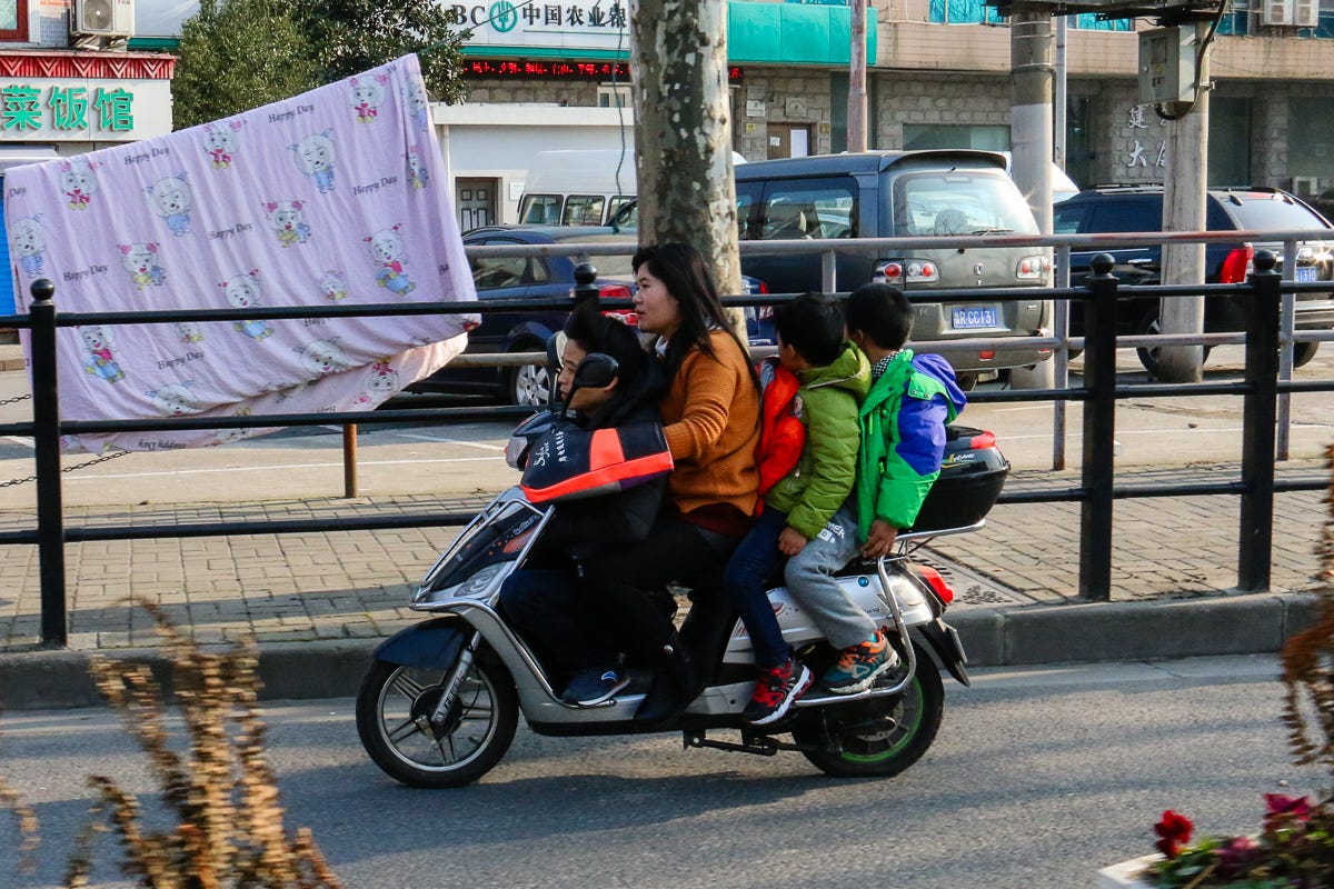 A woman and three children riding a single moped in Shanghai, with a laundry line and cartoon bedsheet in the background. Urban family life meets quirky transport and daily routine.