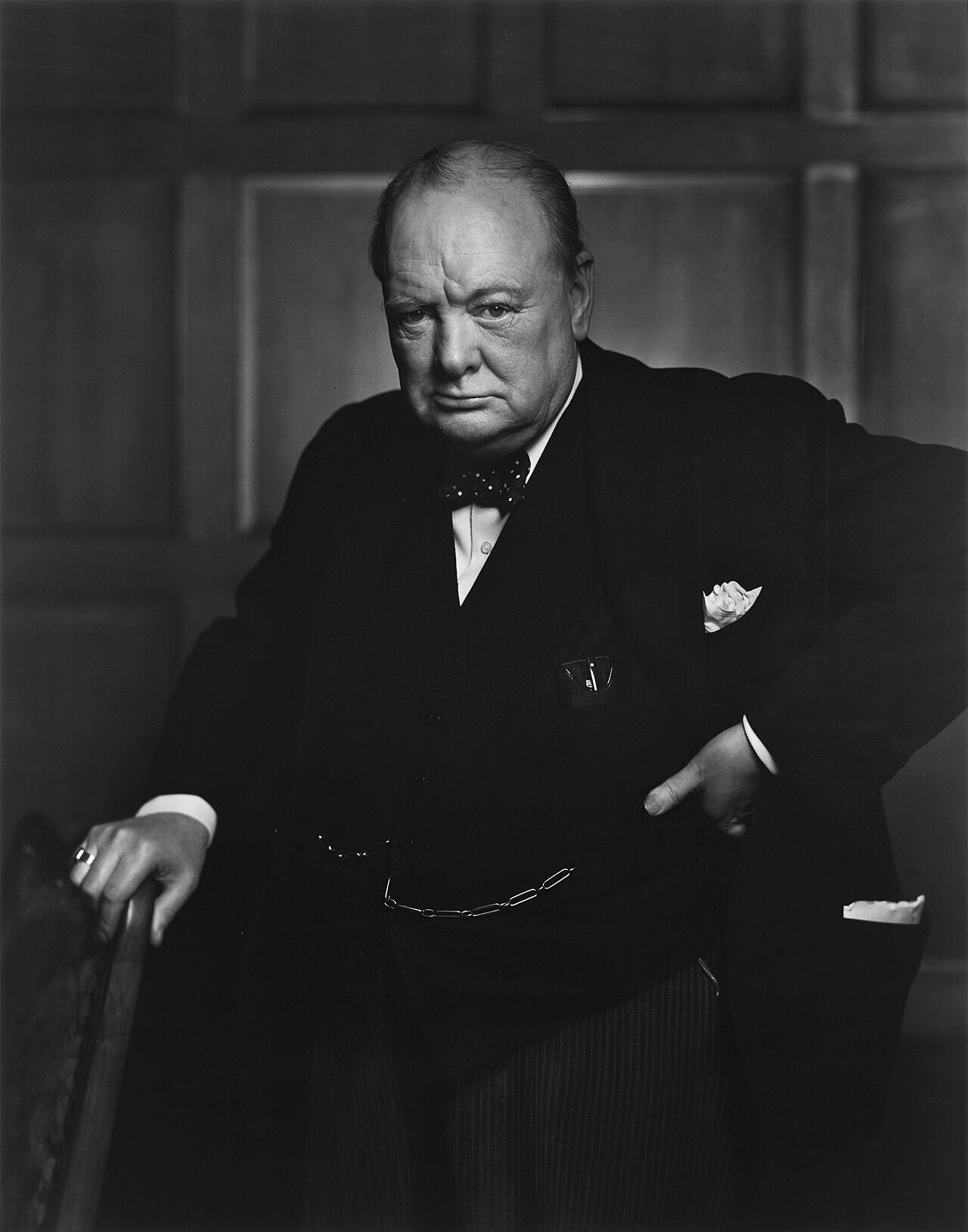 Three-quarter length portrait photograph of Churchill, 67, wearing a suit, standing and holding into the back of a chair