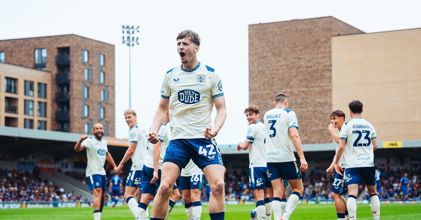 Huddersfield Town’s George Sebine celebrates a goal in front of the away fans at Wimbeldon’s stadium. His teammates can be seen joining in the revelry in the background.