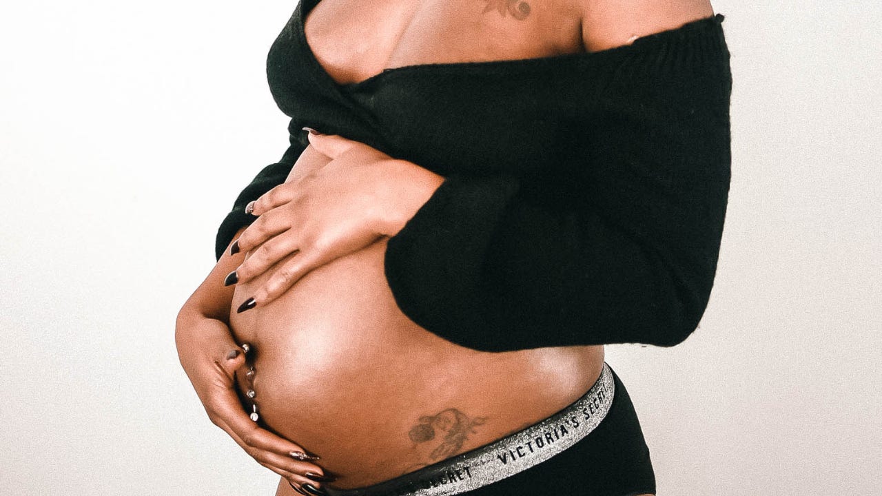 Pregnant Black woman wearing a black off-the-shoulder sweater and black bottoms, gently cradling her baby bump with both hands against a neutral background.