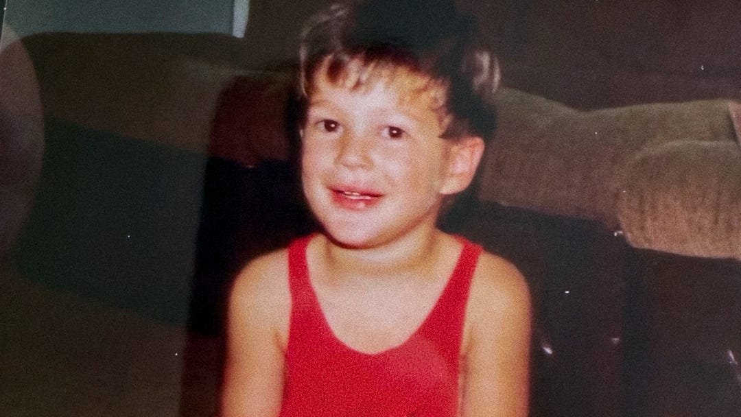Vintage scanned image shows a young child with short brown hair wearing a bright red tank top, smiling directly at the camera. The child is sitting indoors with a brown couch visible in the background.