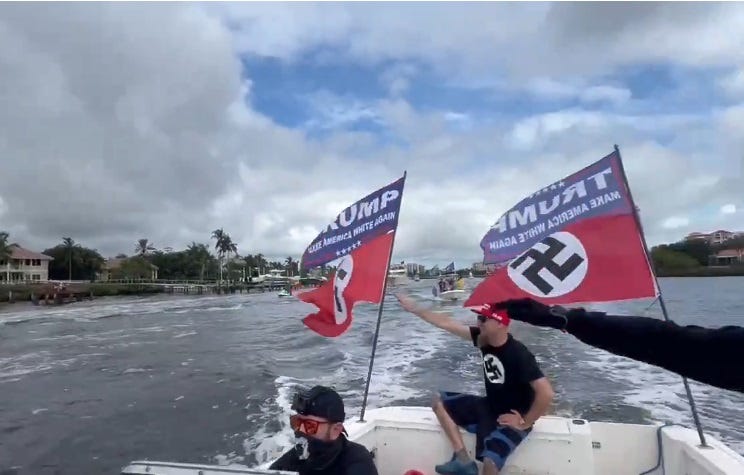 Video screenshot of three men in a small boat that's flying Swastika flags beneath Trump flags. Two of the men have their arms raised in a Nazi salute. All wear black, and one man has a big swastika emblem printed on his T-shirt. Video screenshot of three men in a small boat that's flying Swastika flags beneath Trump flags. Two of the men have their arms raised in a Nazi salute. All wear black, and one man has a big swastika emblem printed on his T-shirt.