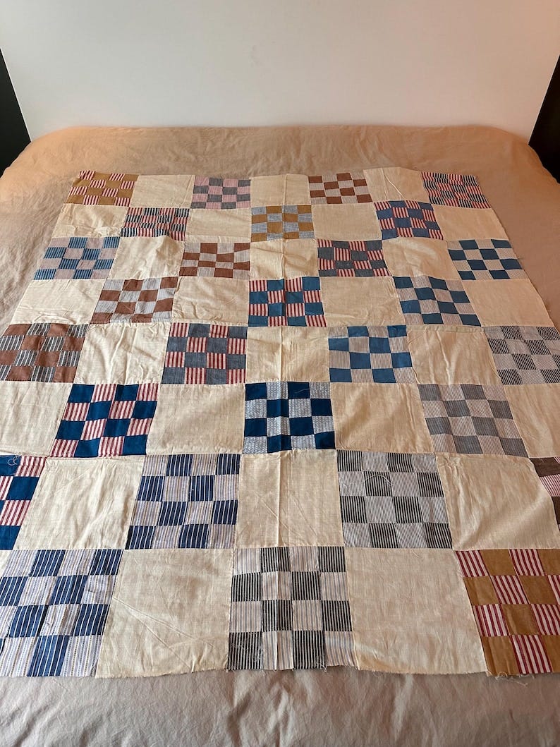 May include: A patchwork quilt featuring a grid of alternating solid off-white fabric squares and squares with checkered patterns in various colors including blue, brown, and red-and-white stripes. The textile is constructed from multiple stitched fabric blocks.