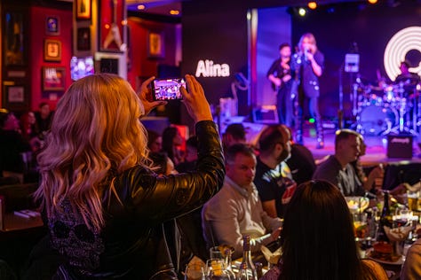Alina Crișan & Keo doi artiști cântă împreună pe scenă, față în față, fiecare ținând câte un microfon. Artista poartă o ținută neagră, iar artistul poartă o geacă din piele. Fundalul scenei are elemente grafice luminoase în tonuri de verde și galben, iar atmosfera este specifică unui concert live.