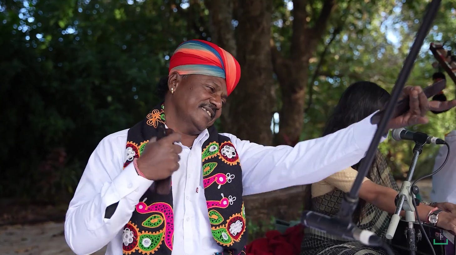 A man playing a khartal under trees, with a woman singing in the background