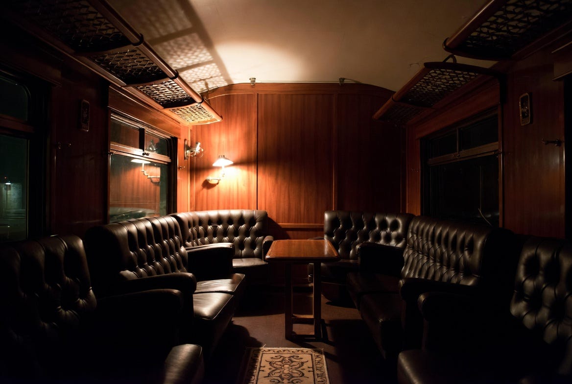 Dark interior of a private 1910s railway car at night, dim lighting, leather chairs arranged around a central table, heavy wood paneling